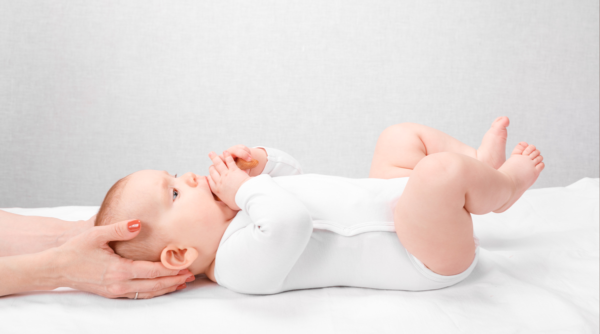 Ein Baby liegt in weißem Strampler auf dem Rücken und eine Frau hält den Kopf des Kindes